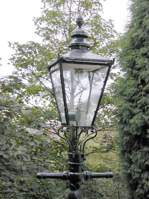 Free Stock Photo: Old cast iron Victorian gas lamp with the traditional lantern style glass on a street with leafy green trees, close up view of the streetlight