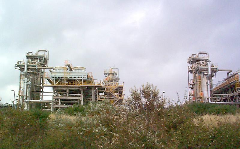 Free Stock Photo: Infrastructure of an industrial plant or factory with external pipework viewed over bushes against a cloudy sky