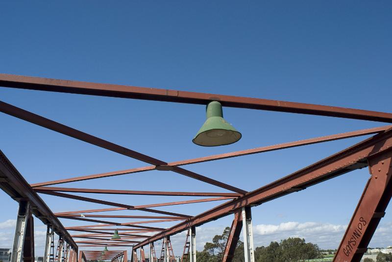Free Stock Photo: Overhead rusty girders of a bridge structure with an electric light with shade in an open framework against a blue sky
