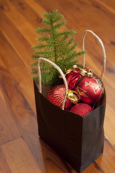 Free Stock Photo: Recyclable paper carrier bag filled with colorful red with Christmas decorations and a small green pine tree to decorate on a wooden floor