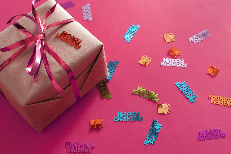 Free Stock Photo: Over View of Gift Wrapped in Brown Paper and Pink Foil Ribbon Surrounded by Birthday Themed Confetti on Hot Pink Background