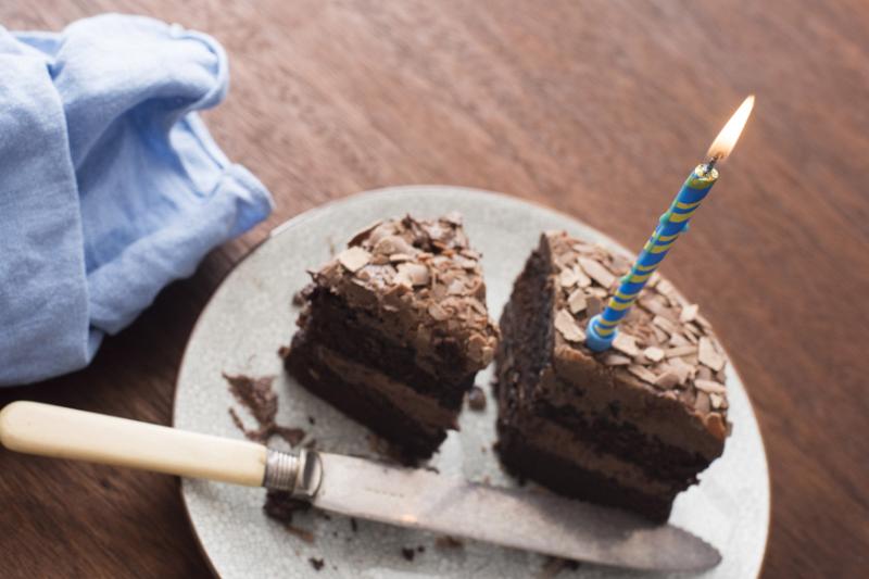 Free Stock Photo: Chocolate cake with lit blue candle on white plate next to dull knife and cyan cloth napkin atop wooden table