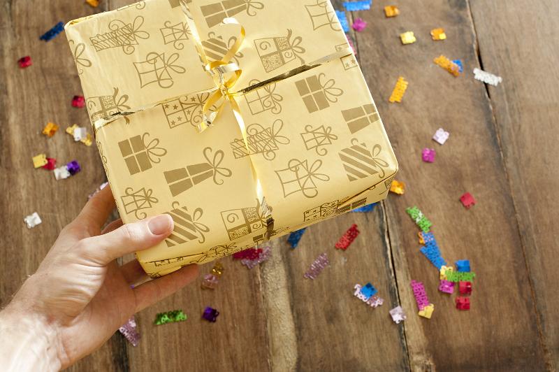 Free Stock Photo: Man giving a gift at a party holding a decorative gift-wrapped box over a wooden table strewn with colorful confetti, close up view of his hand