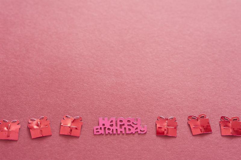 Free Stock Photo: Red Metallic Birthday Confetti Shaped Like Presents with Birthday Message Forming Border Along Bottom of Frame on Red Background with Copy Space