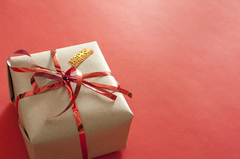 Free Stock Photo: Gift Wrapped in Brown Kraft Paper and Shiny Red Ribbon Tied in Bow with Happy Birthday Confetti on Red Background with Copy Space