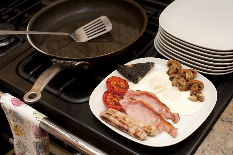 Free Stock Photo: Making breakfast with a stack of plates alongside a range with one breakfast of fried eggs, bacon, mushrooms, tomato and plack pudding already served on a plate