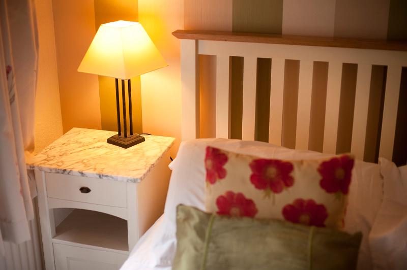 Free Stock Photo: Bedside table and lamp alongside a bed with a simple slatted wooden headboard with floral cushions on the counterpane