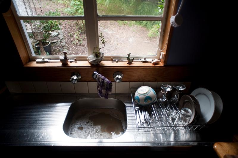Free Stock Photo: View of a stainles steel kitchen sink with dirty water and washing up on the side in the shadows in front of a window overlooking a back garden