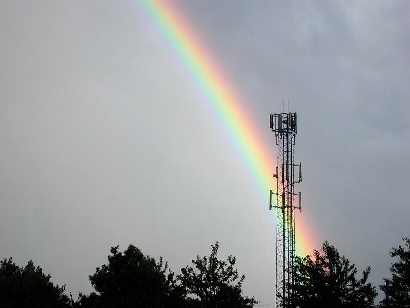 Free Stock Photo: Colorful rainbow of refracted light on water droplets arcing through the sky behind a telecommunications tower on a wet rainy day