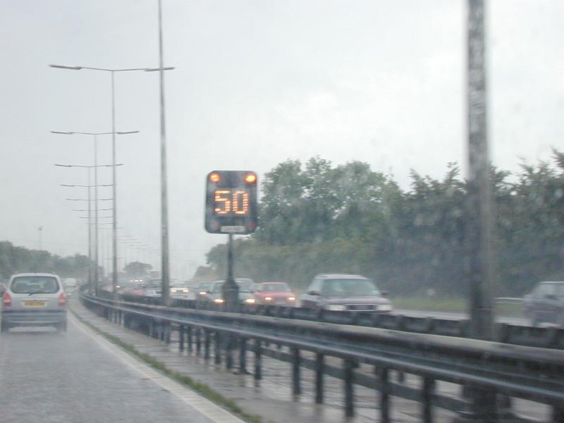 Free Stock Photo: An illuminated 50 speed sign on a motorway in the center behind a safety barrier on a rainy day with traffic
