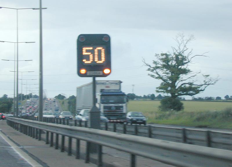 Free Stock Photo: Illuminated digital motorway traffic speed sign in the centre of a multi lane highway with trucks and cars on a rainy day