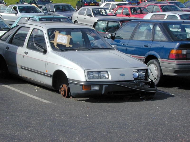 Free Stock Photo: Old silver sedan car parked in a parking lot with a missing front tyre with a notice taped to its windscreen as the axle rests on the tarmac
