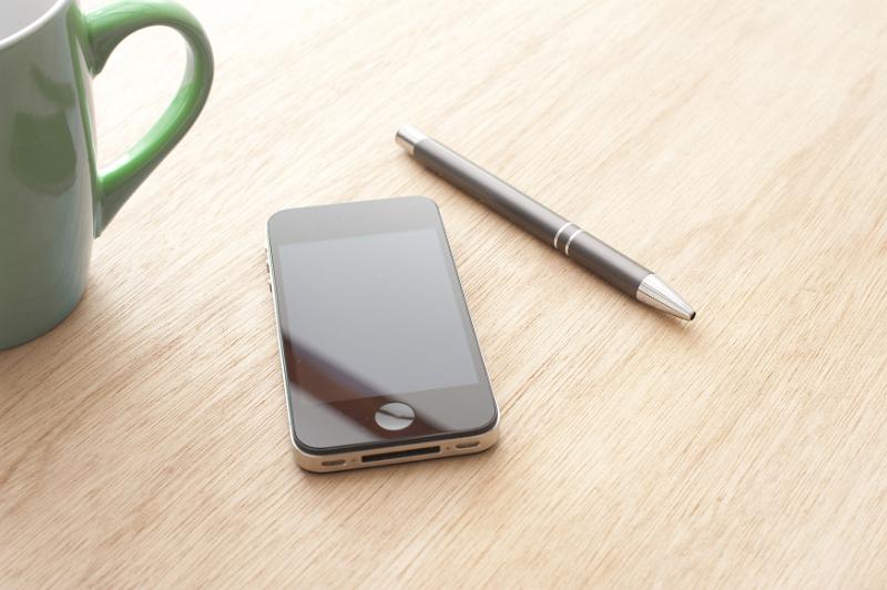 Free Stock Photo: Modern mobile phone with a blank black touchscreen lying on an office desk with a ballpoint pen and mug of coffee