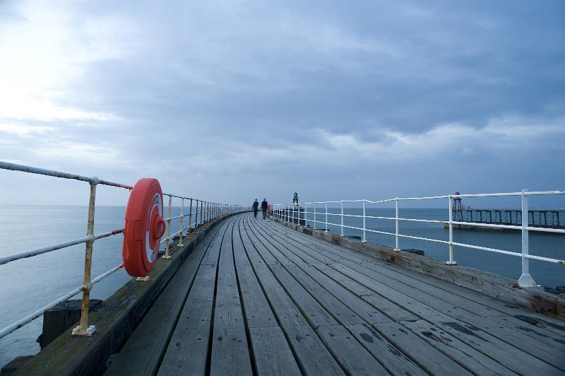 Free Stock Photo: Orange lifering mounted on the railings of a wooden pier stretching out into the ocean with pedestrians visible in the distance